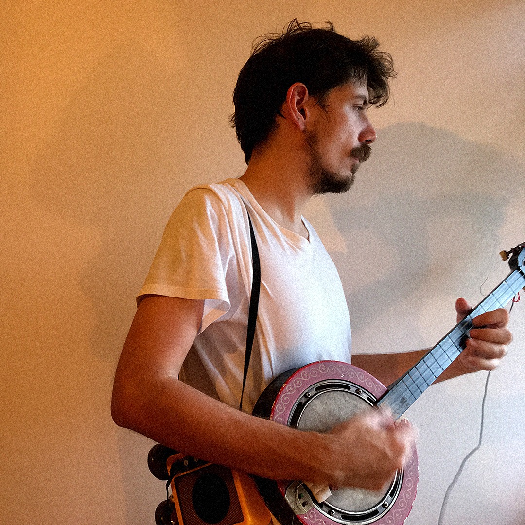 Fotografia colorida. Bruno está com o corpo virado para o lado esquerdo segurando um bandolim. A mão esquerda de Bruno está em movimento tocando o bandolim e a mão direita apoiada sob as cordas. Bruno tem cabelos curtos e escuros, barba e bigode pretos e orelhas grandes. Ao fundo vê-se uma parede bege, com a sombra dele refletindo sobre ela.