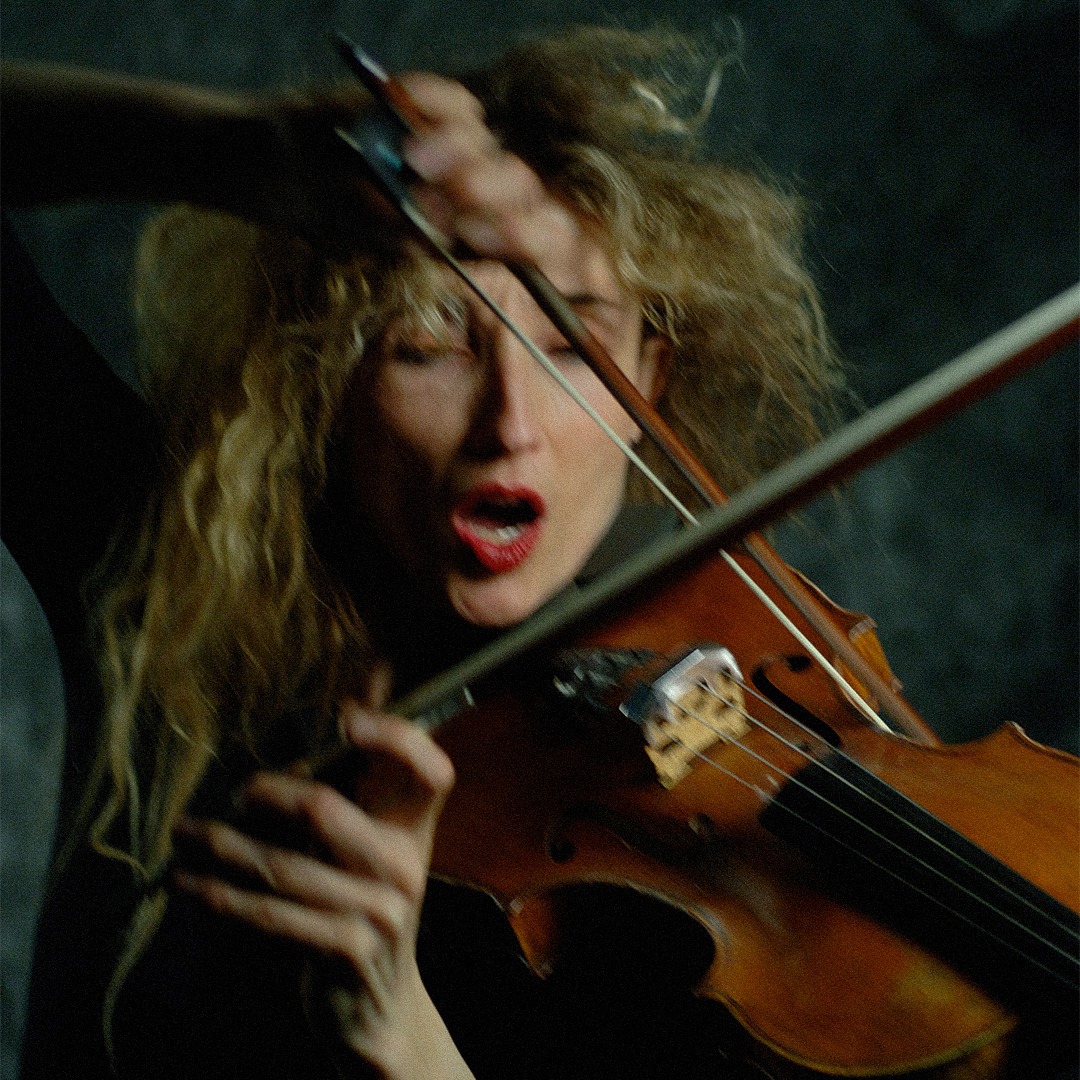 Fotografia colorida. Charlotte é vista de frente tocando uma viola. Ela segura a viola no queixo com um arco em cada mão. Vê-se a mão esquerda sob a testa, bagunçando os cabelos dela. A boca dela boca está aberta com batom vermelho intenso e seus olhos estão fechados.