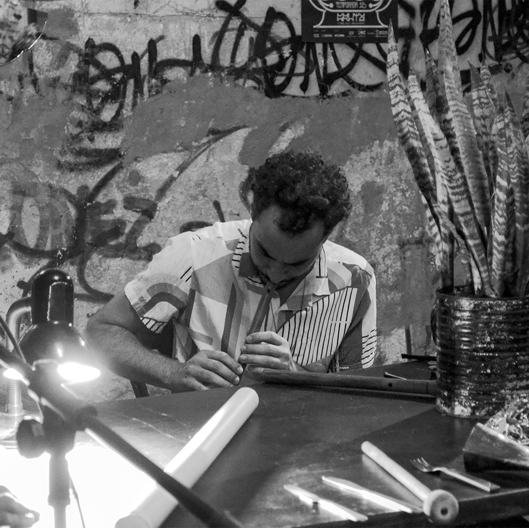 Fotografia em preto e branco. Luiz está sentado em frente a uma mesa. Há sob a mesa uma lata, utilizada como vaso, com a planta espada de São Jorge ao lado direito e uma luminária e uma lata de cerveja do lado esquerdo. Luiz está sentado ao centro e toca uma flauta. Ele segura a flauta com as duas mãos e está com a cabeça virada para baixo. Luiz tem cabelos curtos, escuros e encaracolados com entradas nas laterais da testa. Usa uma camisa estampada com mangas curtas.